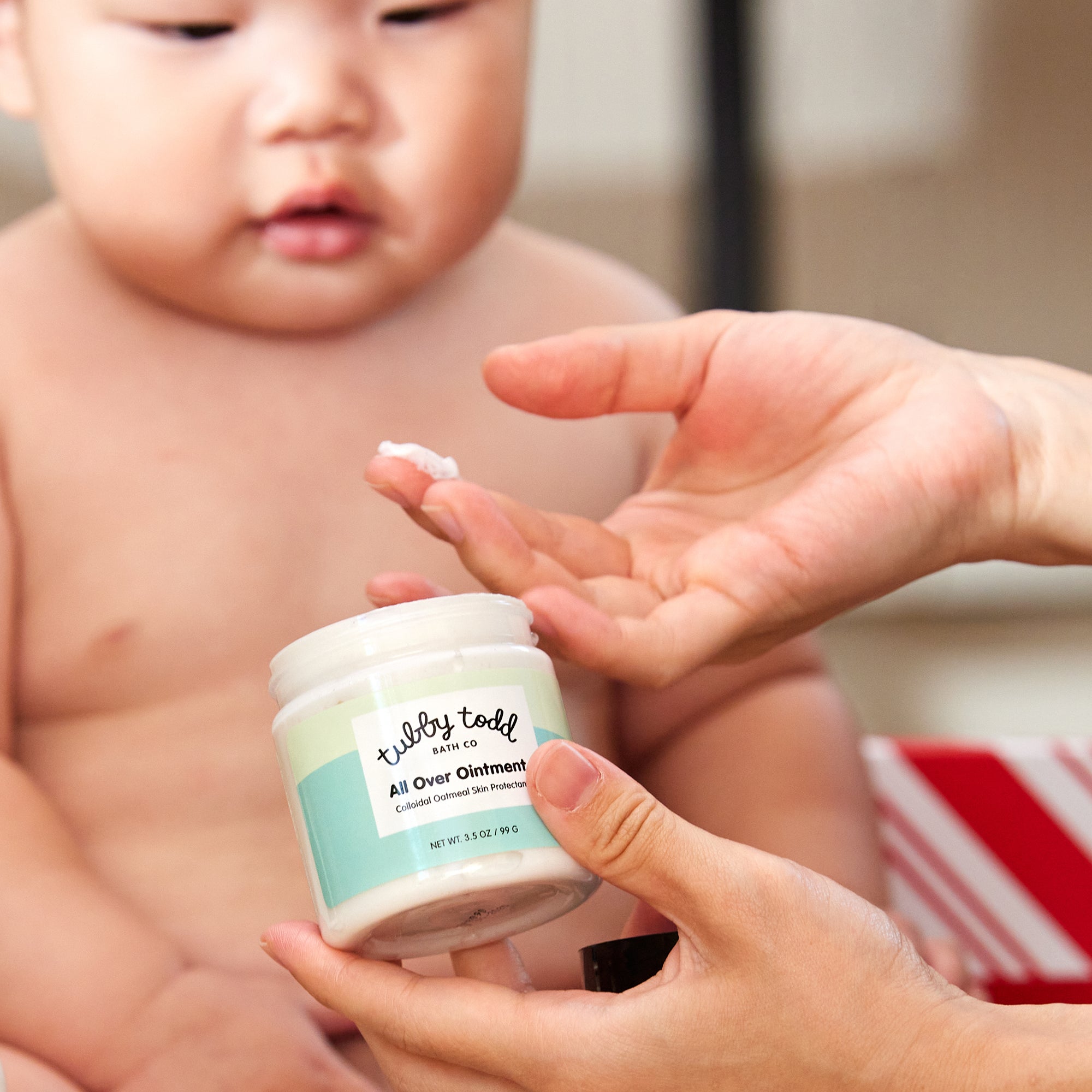 Hand holding the 3.5oz All Over Ointment and scooping out the cream on the finger, and the baby boy seeing that.